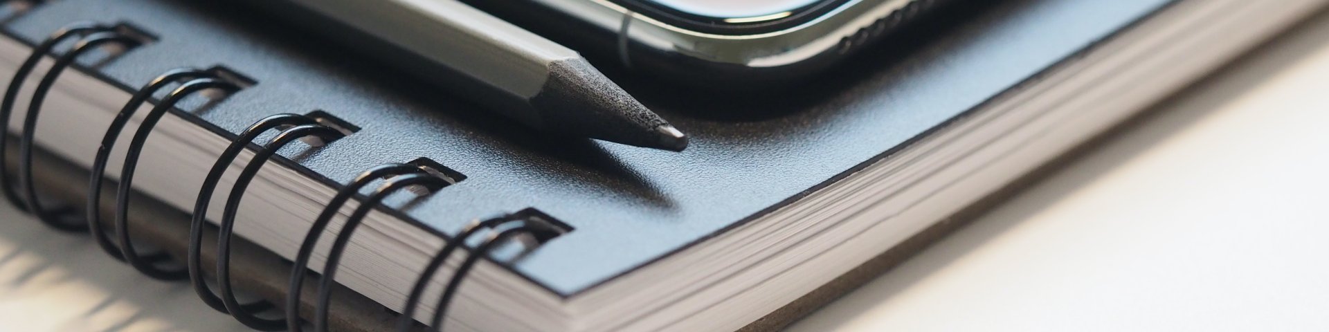 Closeup Photo of Black Smartphone Near Black and Grey Pencil on Black Spiral Notebook